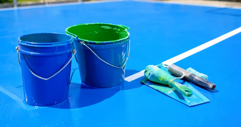 Hai thùng sơn sân thể thao màu xanh dương và xanh lá, cây lăn và bàn gạt đặt trên mặt sân acrylic xanh có vạch trắng